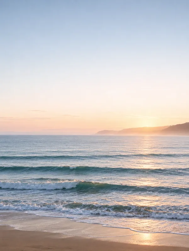 Rolling turquoise waves catch warm morning light under a pale sky, with the coastline fading into the distance.