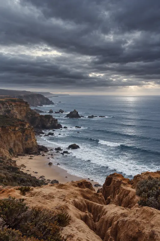 Stormy coastal cliffs of rust-coloured rock above a deep blue sea with breaking waves beneath dark clouds.