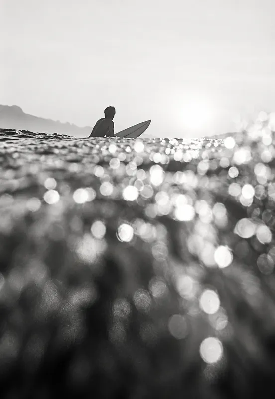 Black-and-white photo of a surfer silhouetted against bright sun over a field of sparkling water droplets.