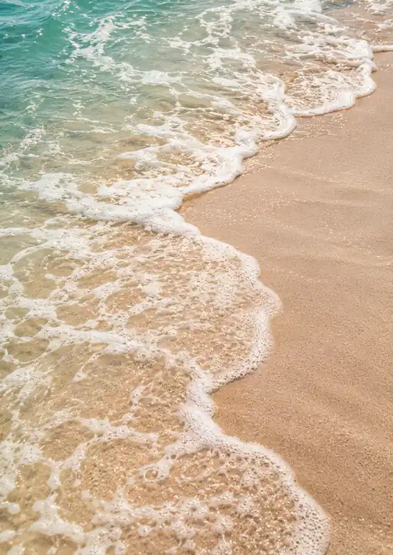 Overhead view of turquoise waves washing over pale sand, leaving curling patterns of white foam.