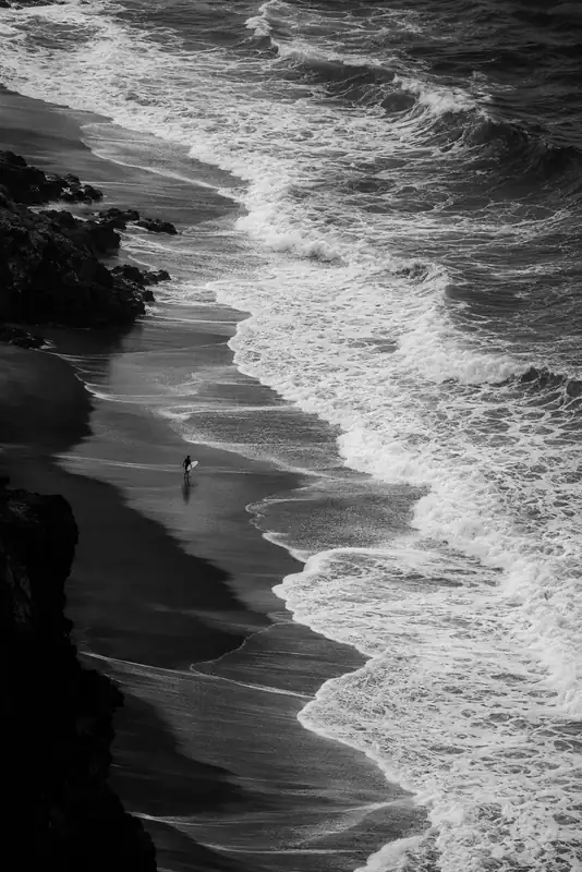 Black-and-white aerial shoreline with rocky edges, churning surf and a tiny figure on the sand.