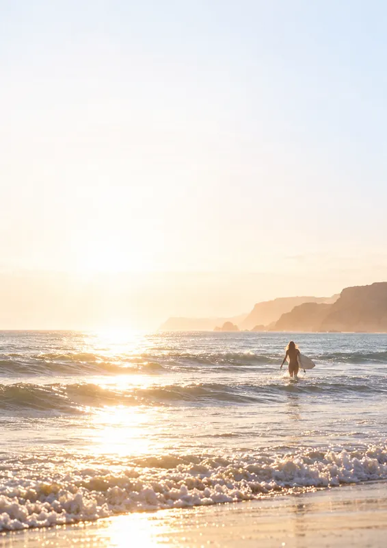 Surfer wading into the sea at sunrise, with a glowing horizon, low cliffs and foamy shallows.