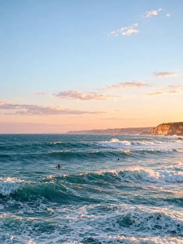 Small surfers in the sea at dusk, with a pastel sky and rolling blue waves across the frame.