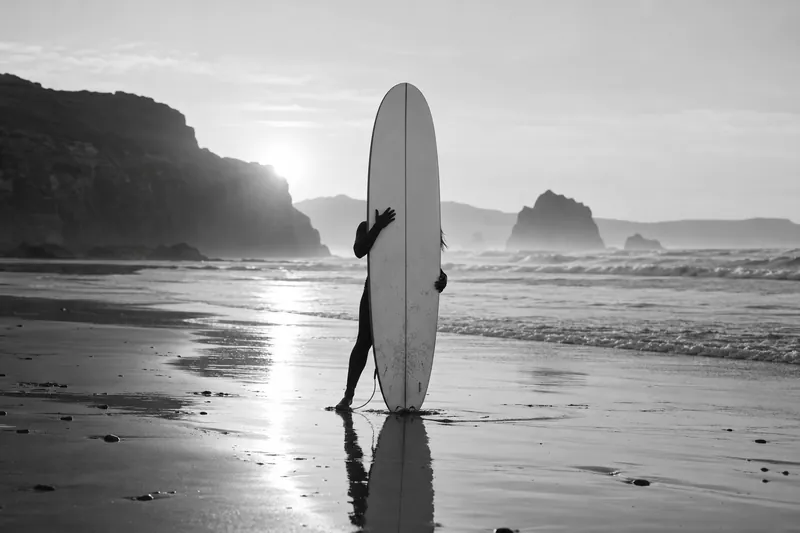 Black-and-white photo of a surfer standing on wet sand, holding a board upright with a bright reflection below.