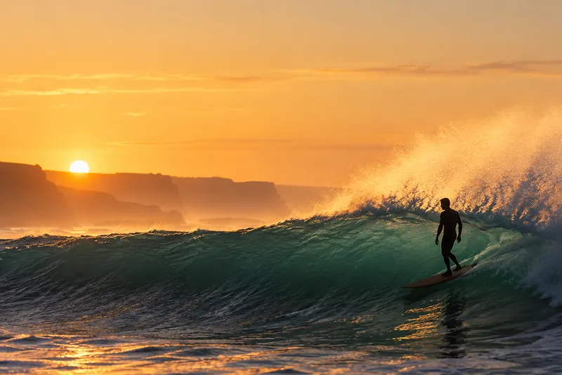 Surfer riding a golden-lit wave at sunset, silhouetted against a bright sky and a cliff line.