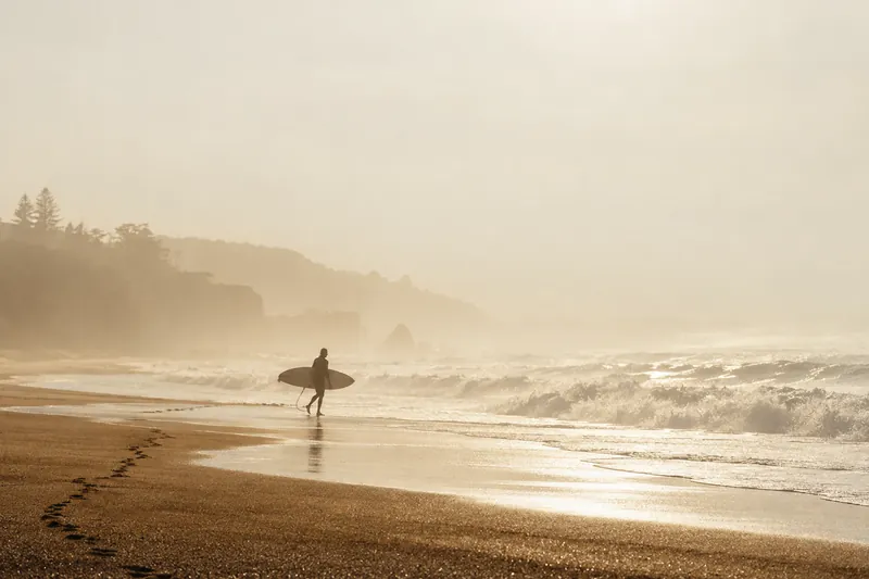 Lone surfer on a misty beach at morning, with footprints across the sand and soft golden haze.