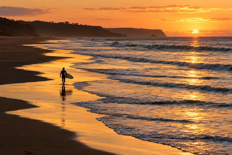 Warm sunset beach with a surfer far off on the sand, golden light reflecting off the wet shore.