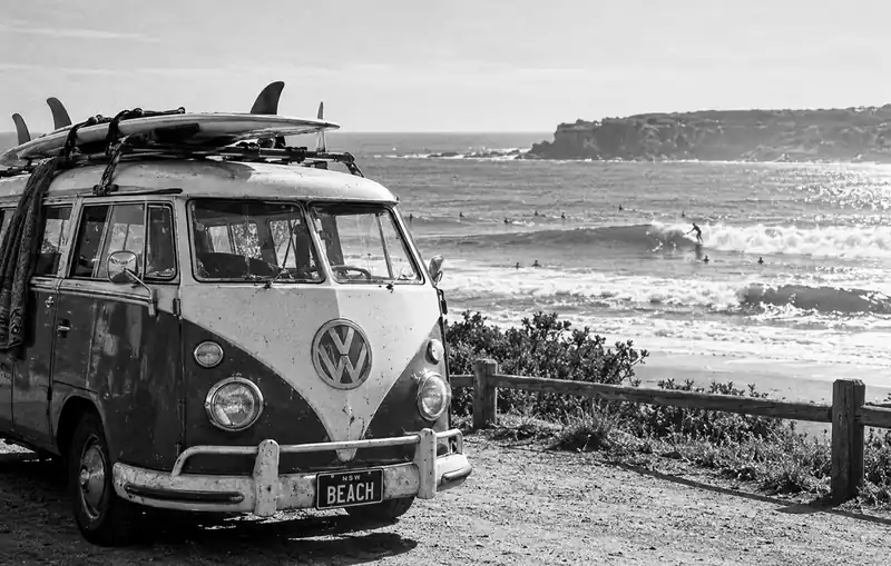Black-and-white photo of a vintage surf van with boards on its roof and a wooden fence along a beach.