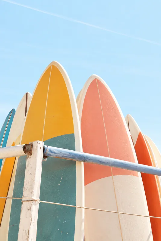Surfboards in teal and coral stand behind a pale wooden fence against a clear blue sky.