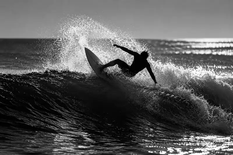 Black-and-white photo of a surfer carving a wave, with dark silhouettes and bright spray.