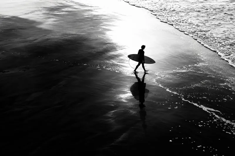 Black-and-white photo of a surfer standing on wet sand with a board, lit from behind by a soft sun.