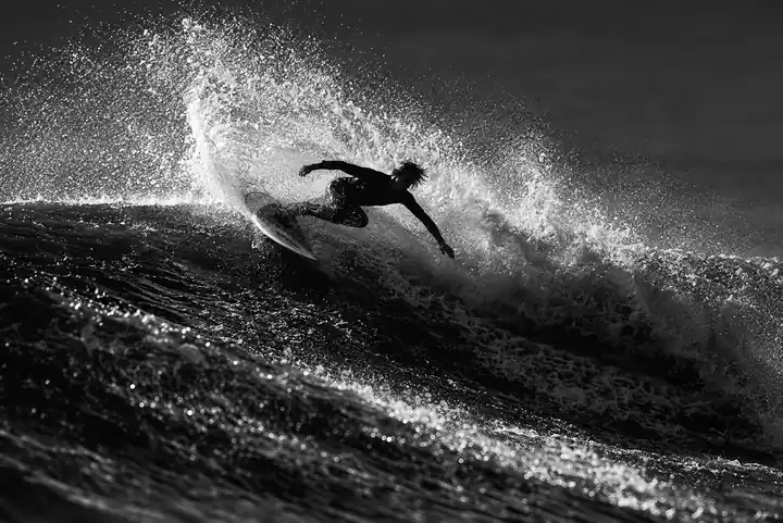 Black-and-white photo of a surfer turning off a wave, with high spray and sharp light across the surface.
