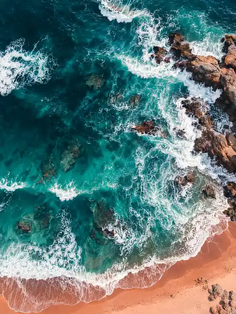 Aerial view of turquoise waves meeting dark rocks and a warm orange sand beach.