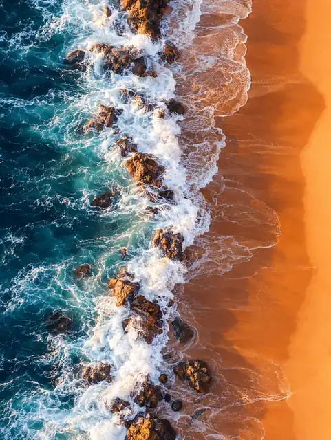 Aerial view of dark rocks and turquoise water meeting a band of warm orange sand.