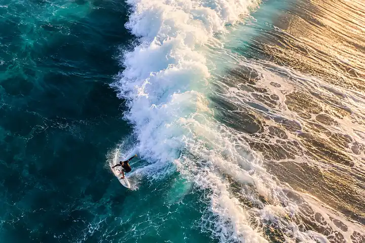 Aerial view of a surfer on a turquoise wave edge with white foam and warm sand below.