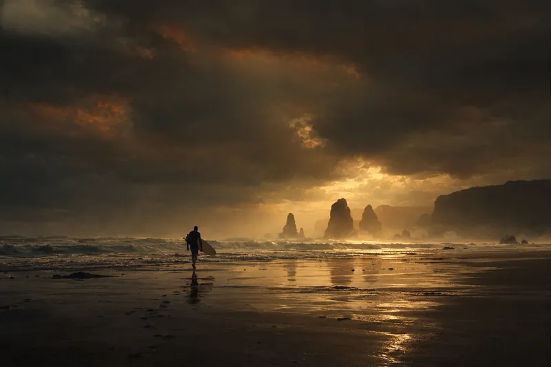 Dark moody beach with a silhouetted surfer and rock stacks in gold light beneath stormy clouds.