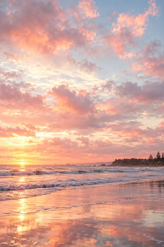 Pink and gold clouds over a wet beach, with a headland in the distance and foam on the sand.